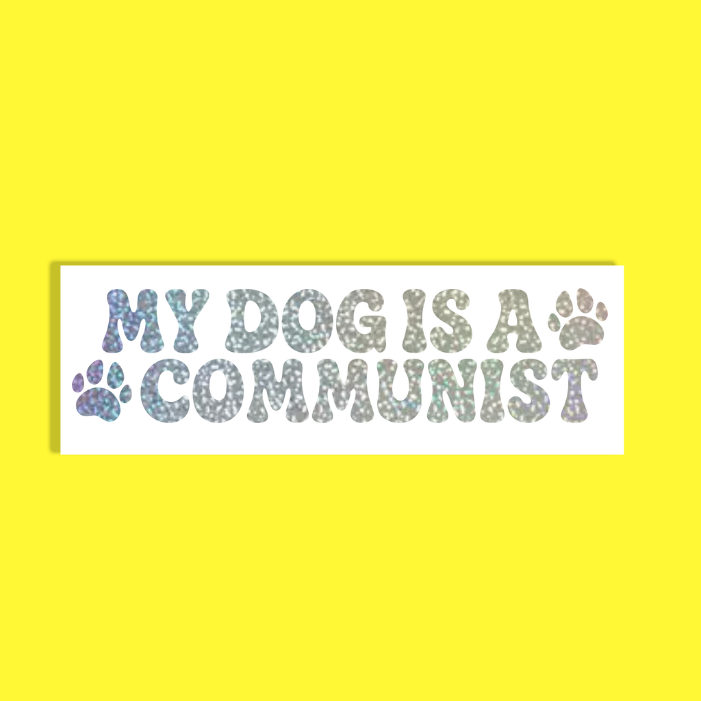 A rectangular white background with the text 'MY DOG IS A COMMUNIST' in a glittery, iridescent font, accented with two paw prints, shown against a solid bright yellow background.
