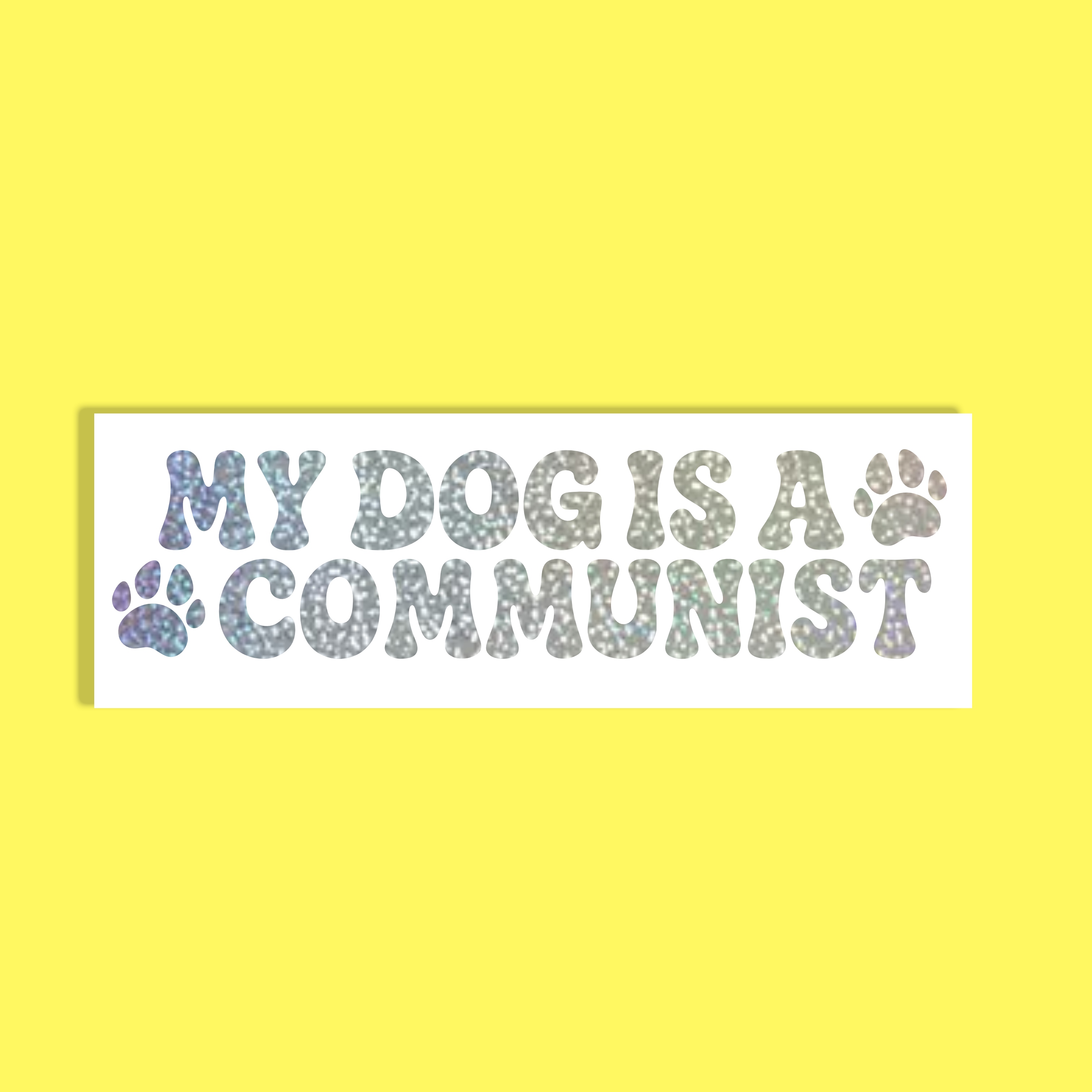A rectangular white background with the text 'MY DOG IS A COMMUNIST' in a glittery, iridescent font, accented with two paw prints, shown against a solid bright yellow background.