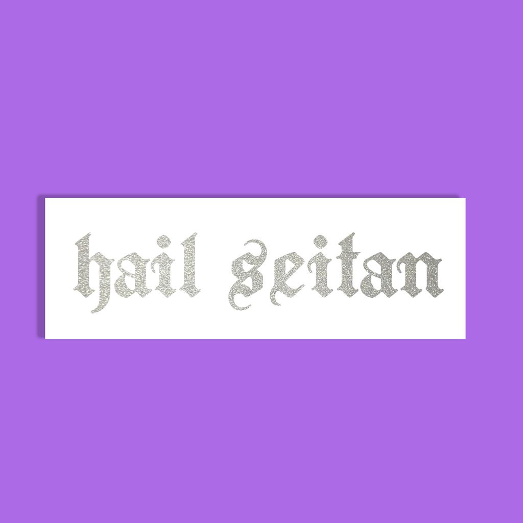 A rectangular white sticker or label with the phrase 'hail seitan' in a glittery silver gothic font, centered on a solid purple background.