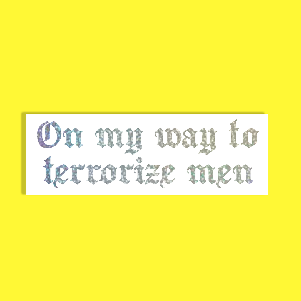 A rectangular white background with the phrase 'On my way to terrorize men' in a decorative, textured silver holographic gothic font, shown against a solid bright yellow background.