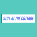 A white rectangle with the phrase 'STILL AT THE COTTAGE' in large blue-purple holographic lettering, centered on a solid teal background.