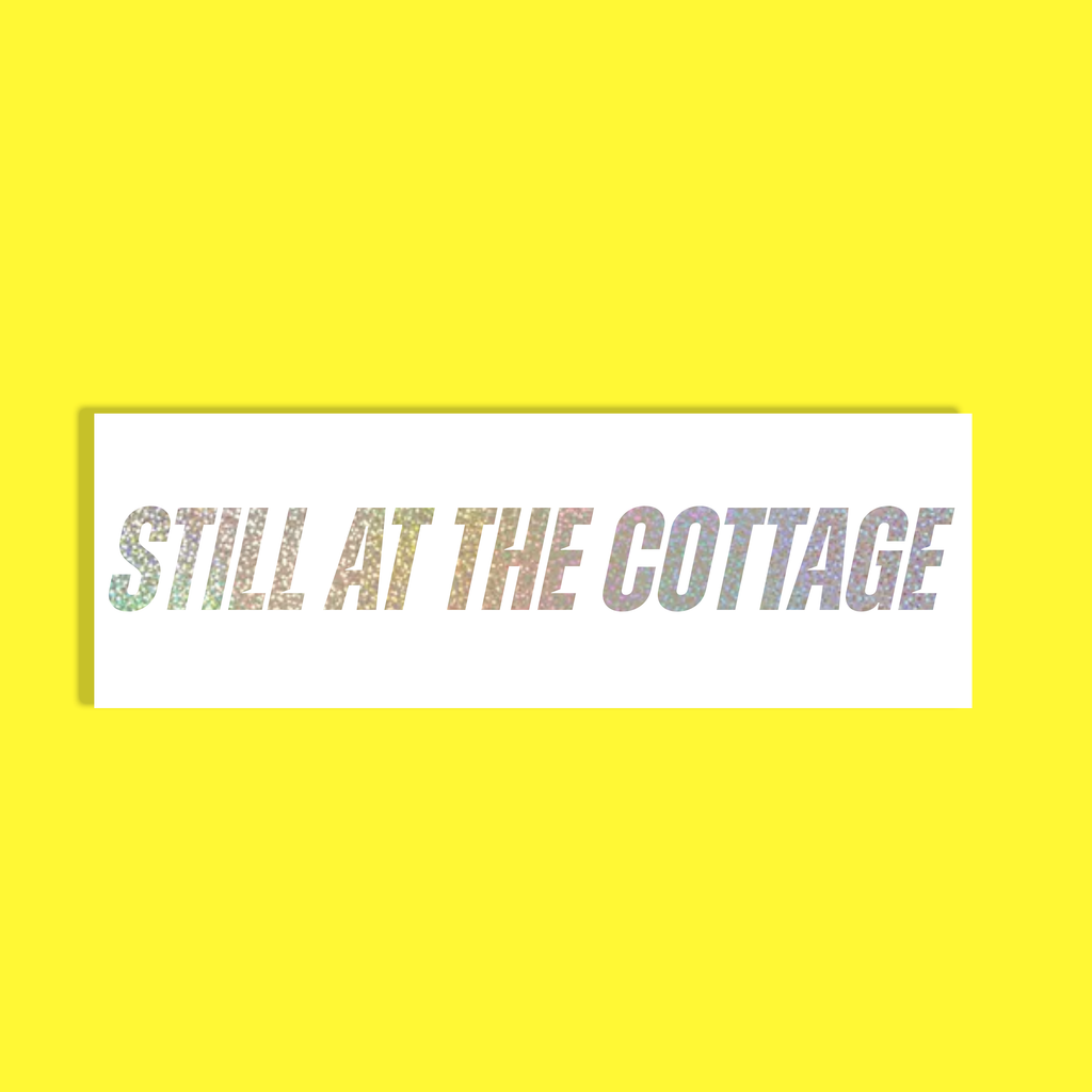 A rectangular white background with the phrase 'STILL AT THE COTTAGE' in silver glittery holographic letters, centered on a solid bright yellow background.